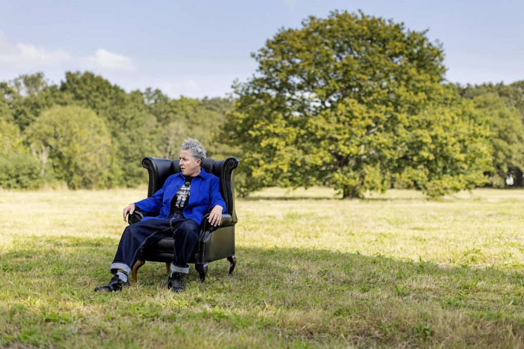 A white-skinned, curly and silver haired human, wearing a royal blue, French-style work coat and jeans, sits in a leather wing-back chair that is in the middle of a field. Behind the chair and the human is framed a large oak tree in the distance and a clear blue sky.